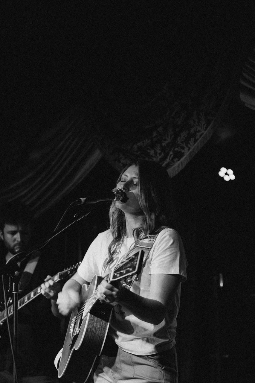 A singer playing acoustic guitar mid-song with eyes closed, black and white